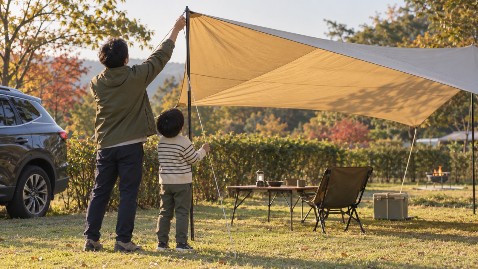 夕方のオートキャンプ場で張ったタープのポールを直す父親と、張り綱を持つ男の子の後ろ姿