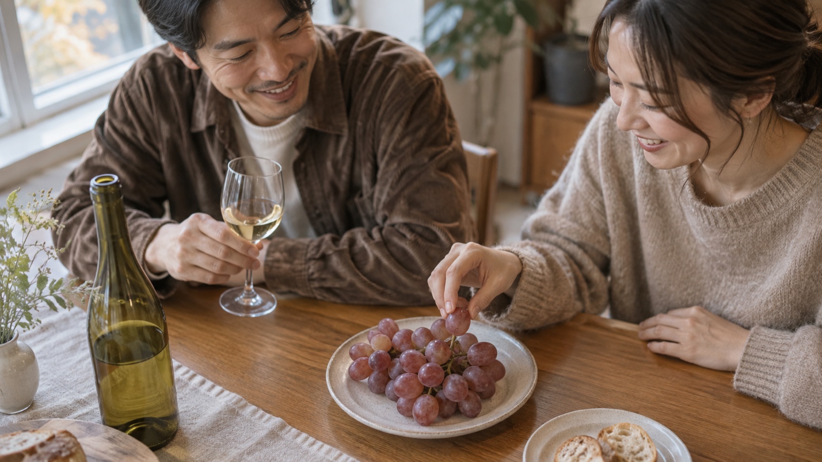 秋の昼下がりの食卓で男女が淡い紫色の生食用ブドウをつまみながら白ワインを味わい、開栓済みボトルが置かれている